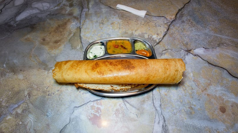 Golden-brown dosa served on a metal plate with four small bowls of chutneys and sambar.